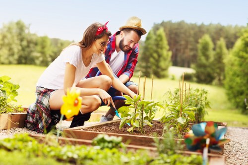 A man and a young girl are working together in a well-maintained garden, tending to a raised vegetable bed filled with young plants. The girl, dressed in a white T-shirt and checkered shorts, wears a red headband with a bow and holds a gardening tool, while the man, wearing a plaid shirt, beige straw hat, and jeans, is assisting her by supporting the plants. Surrounding the vegetable bed is a gravel pathway, and in the foreground, there are several small, green leafy plants. The background features a lush, green lawn with a line of trees and shrubs, indicating a spacious outdoor area with natural sunlight illuminating the scene, suggesting a pleasant, clear day. The garden appears to be part of a private residential outdoor space, showcasing organized planting beds suitable for gardening services offered by Garden Maintenance Penge, including vegetable planting and garden upkeep.