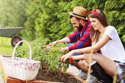 A man and a woman are gardening together in a well-maintained outdoor space, sitting on the ground beside a flower bed. The man, wearing a straw hat and a red and blue plaid shirt, is tending to a small green shrub or young plant, while the woman, dressed in a white T-shirt and plaid skirt with a red headband, assists by holding a garden tool. A white wicker basket with a floral lining is placed nearby on the soil, suggesting they are engaged in planting or tending to the garden. The background features lush green trees and shrubs, indicating a healthy, flourishing garden setting typical of residential outdoor spaces in Penge. The scene is illuminated by natural daylight, with warm, natural tones of soil, greenery, and clothing, highlighting a peaceful gardening activity suited for regular outdoor upkeep services provided by Garden Maintenance Penge, with attention to lawn, plants, and garden bed management.