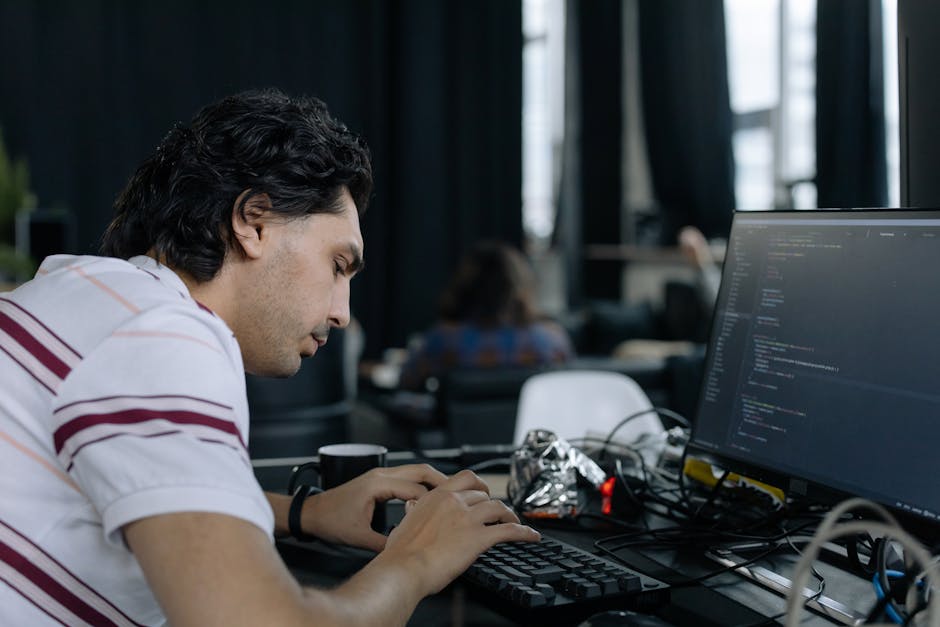 A young man with dark, wavy hair is working at a desk in an indoor office or co-working space, focused on typing on a keyboard connected to a large computer monitor displaying lines of code. The works